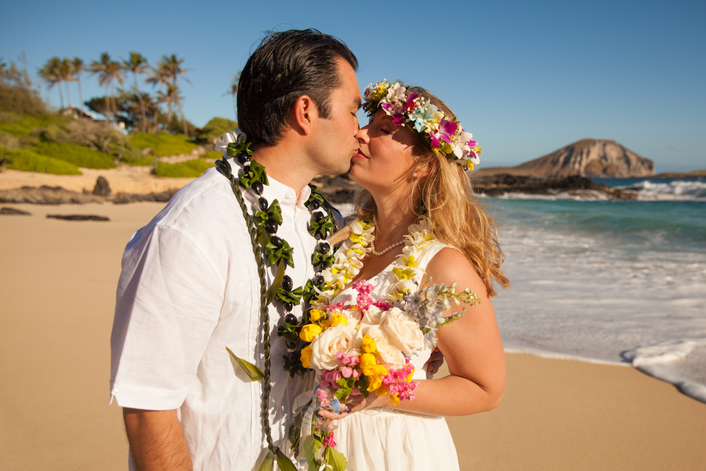 A romantic couple in traditional Hawaiian leis share a kiss on tropical Makapuu Beach with ocean waves and mountains in the background.