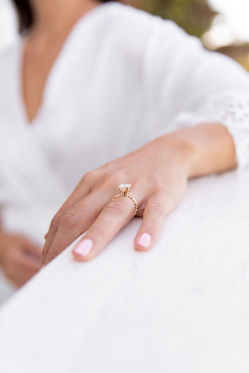 Close-up of a delicate gold engagement ring worn on a woman's hand with white fabric in the background.