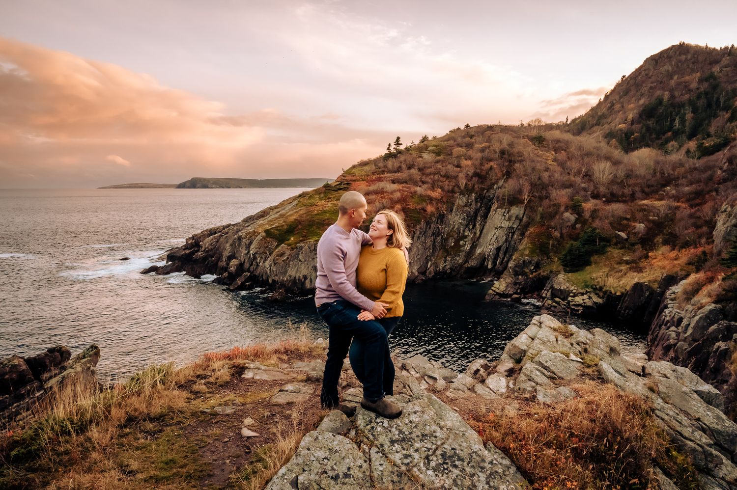 A couple on a rocky shore at sunset, surrounded by warm hues and the rugged beauty of St. John's, Newfoundland.
