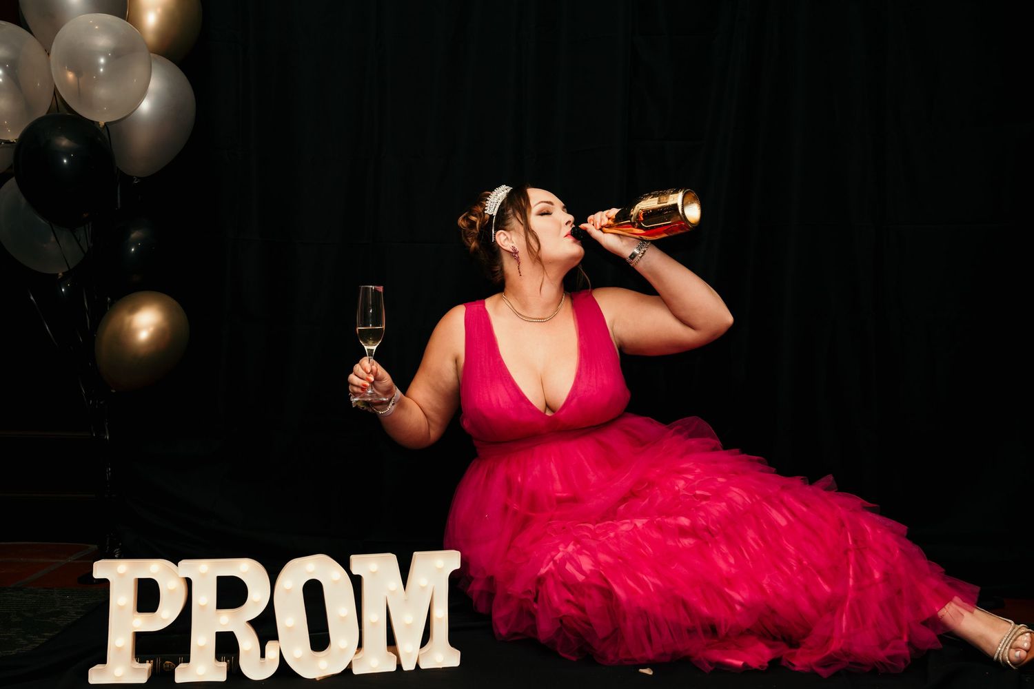 Someone in a bright pink prom dress enjoying drinks and posing with decorative PROM letters.