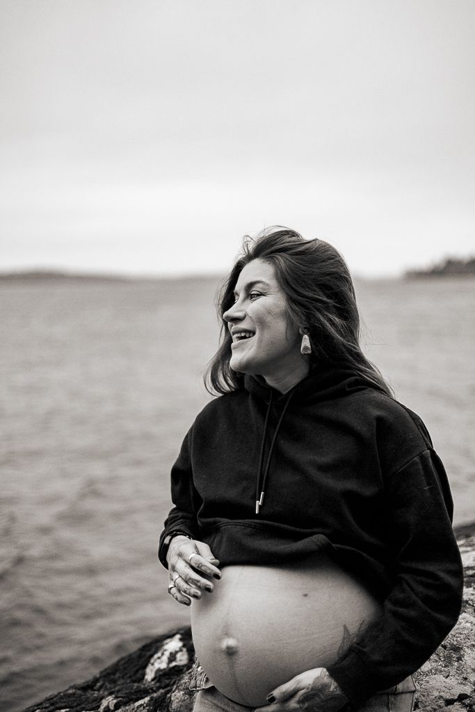 A pregnant person in a grey cardigan stands by a lake at dusk in a moody winter setting.