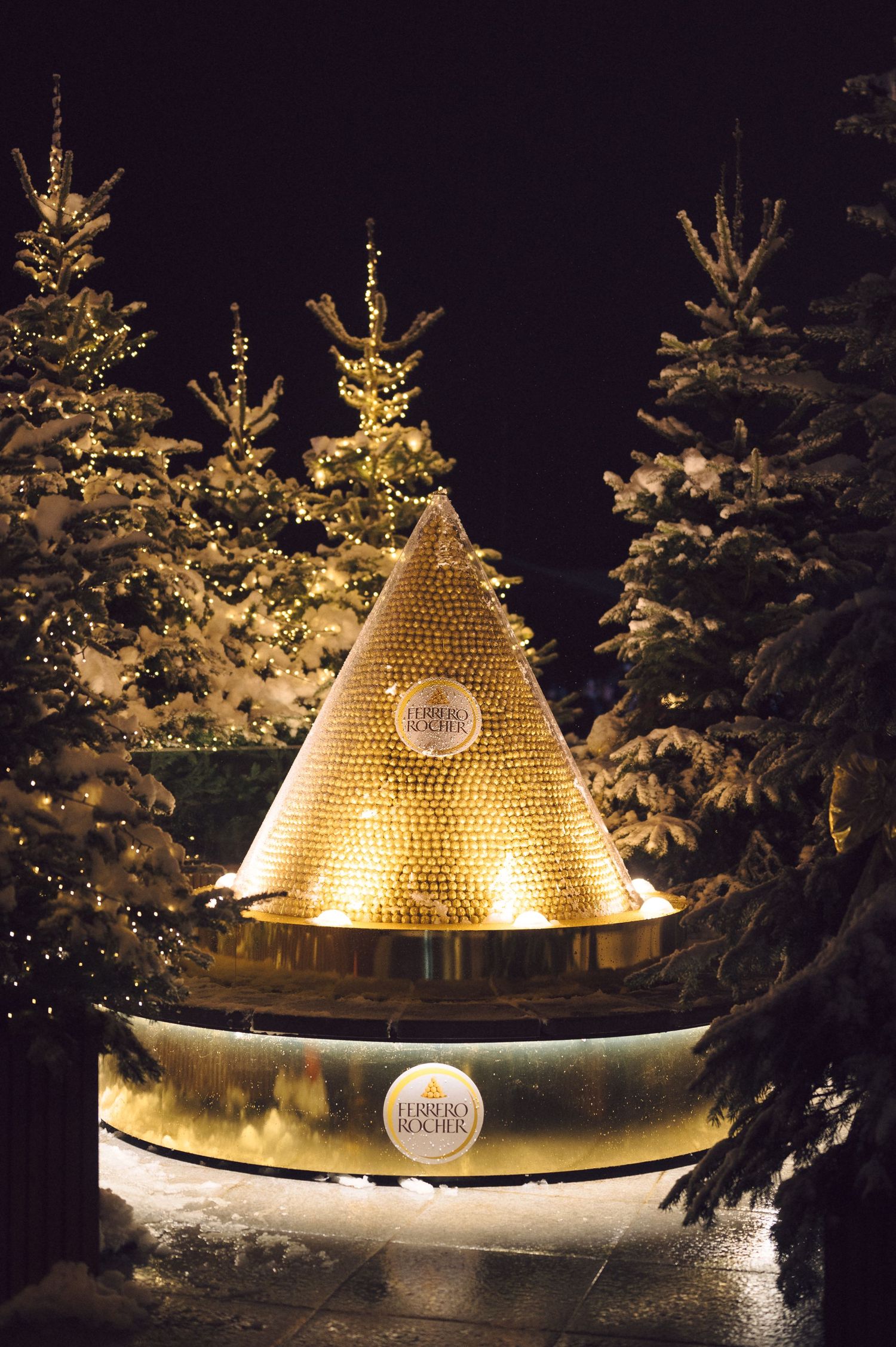 Glowing golden pyramid-shaped art installation surrounded by snow-covered evergreens at night.