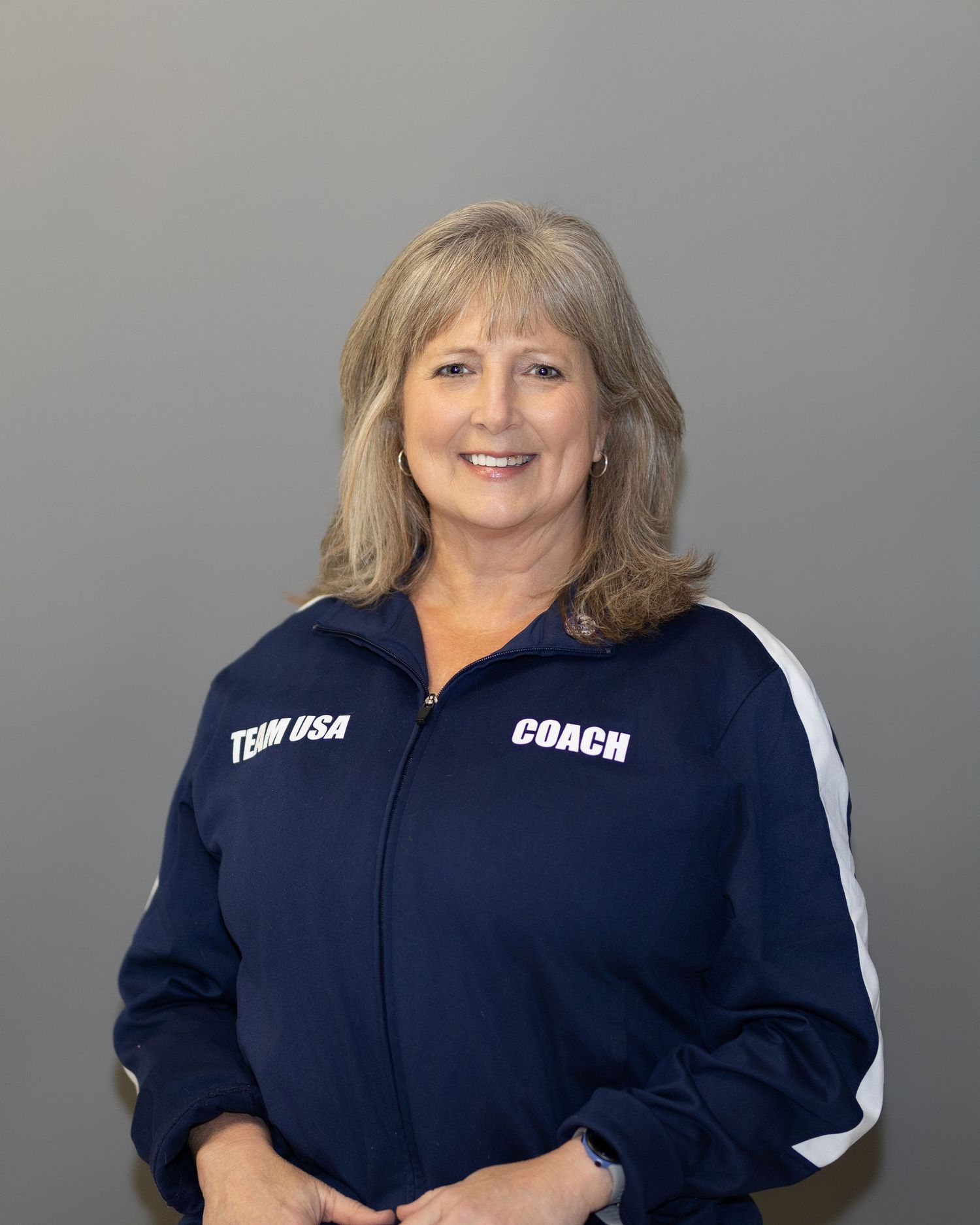 Athletic coach wearing navy blue Team USA jacket poses against gray background.