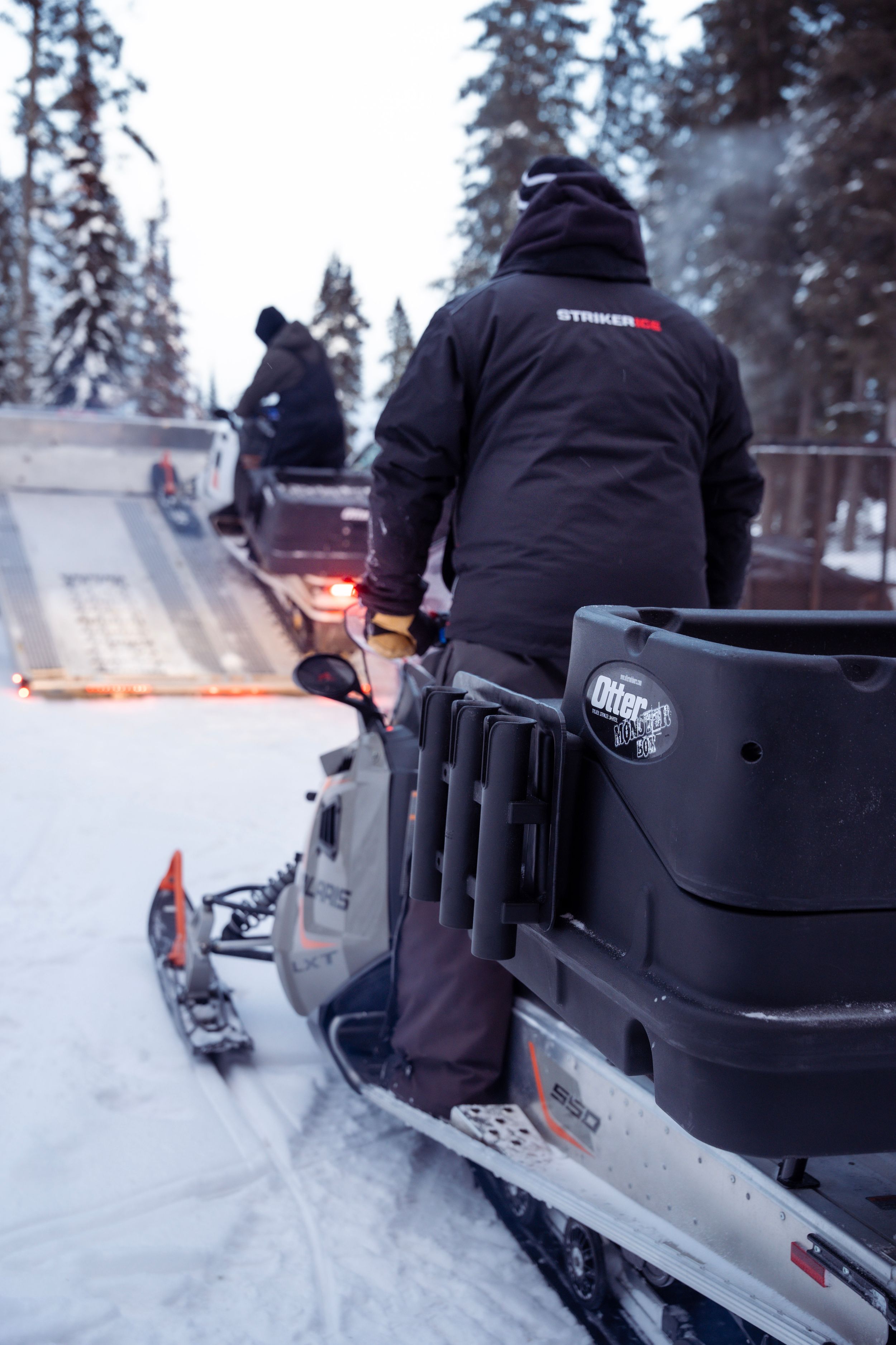 Snowmobiles lined up on snowy trail with riders preparing for departure.