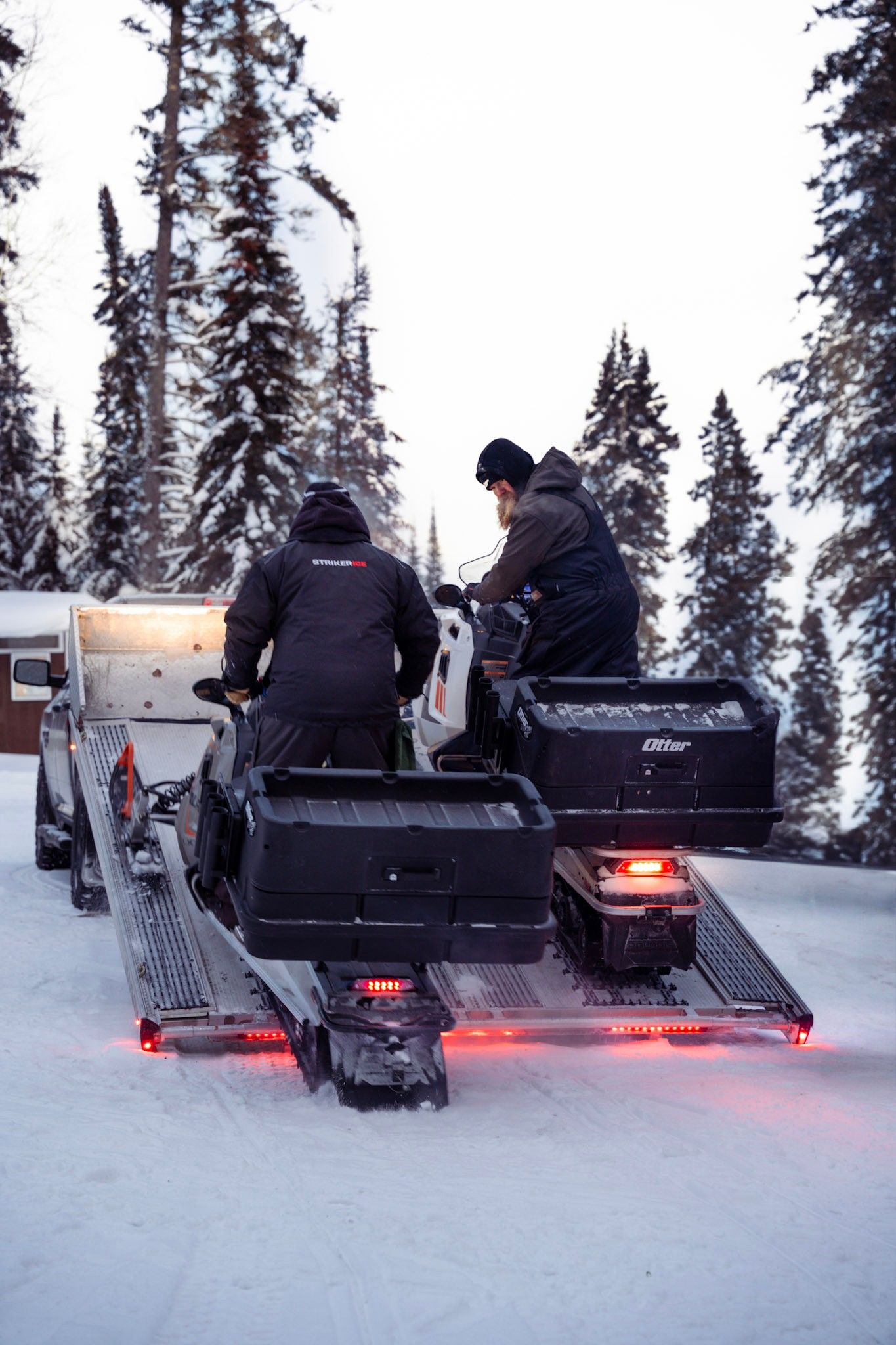 People riding snowmobiles through a snowy forest landscape with red tail lights glowing in the winter dusk.