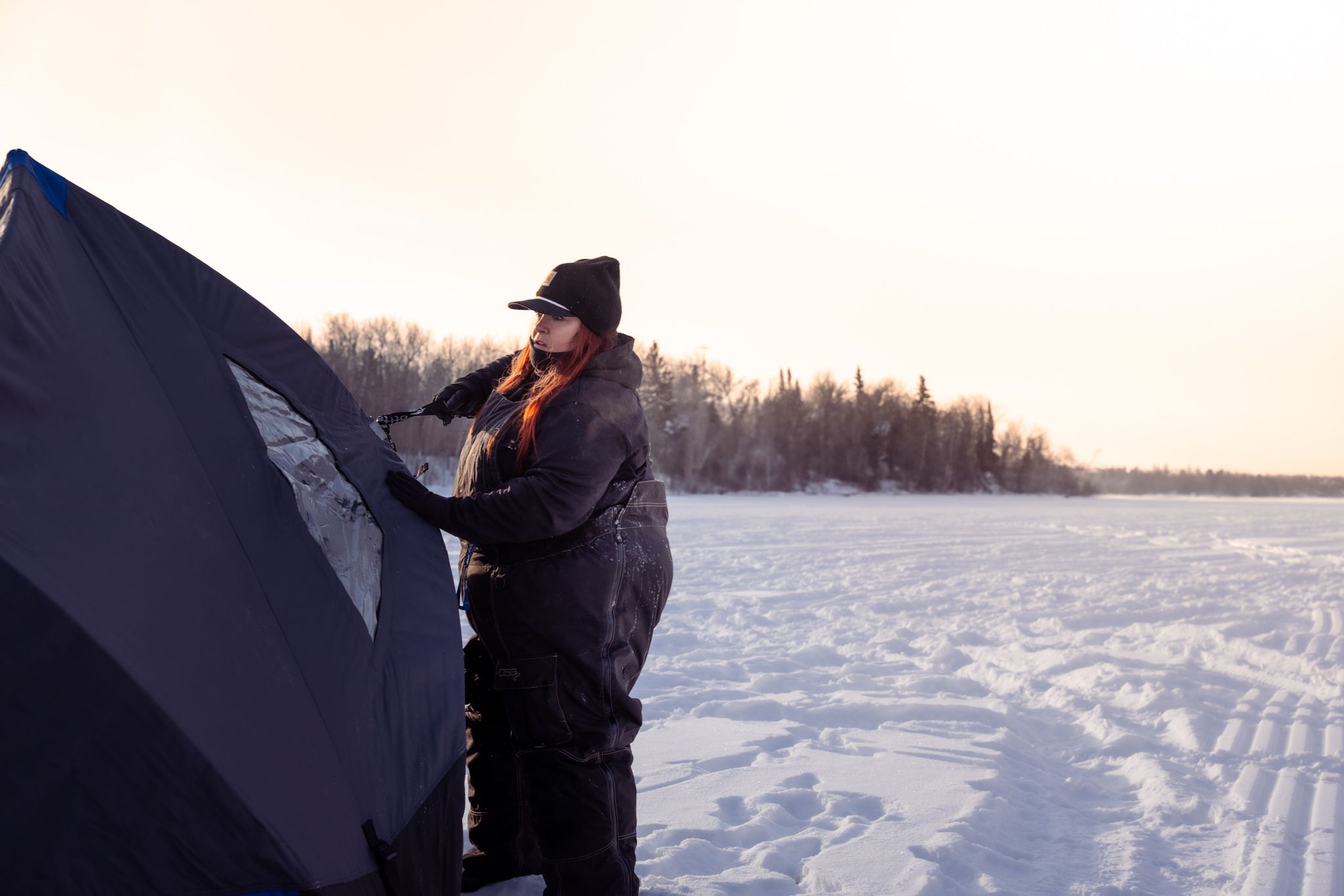 Winter ice fishing scene with someone setting up a dark shelter on a frozen lake at sunset with snow-covered trees in background.