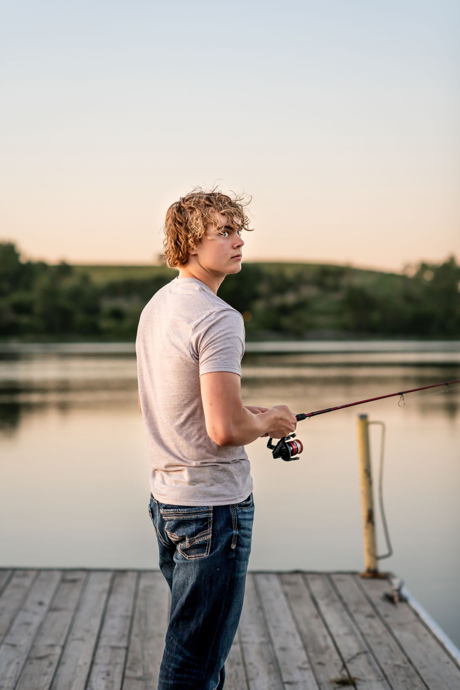 Person fishing on a wooden dock at sunset while wearing casual attire with a lake in the background.