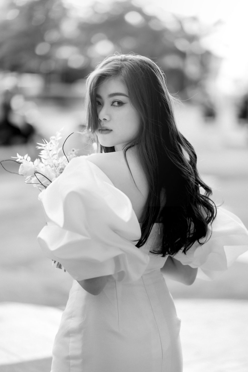 Elegant bride in off-shoulder white gown with puff sleeves holds bouquet in a black and white portrait.