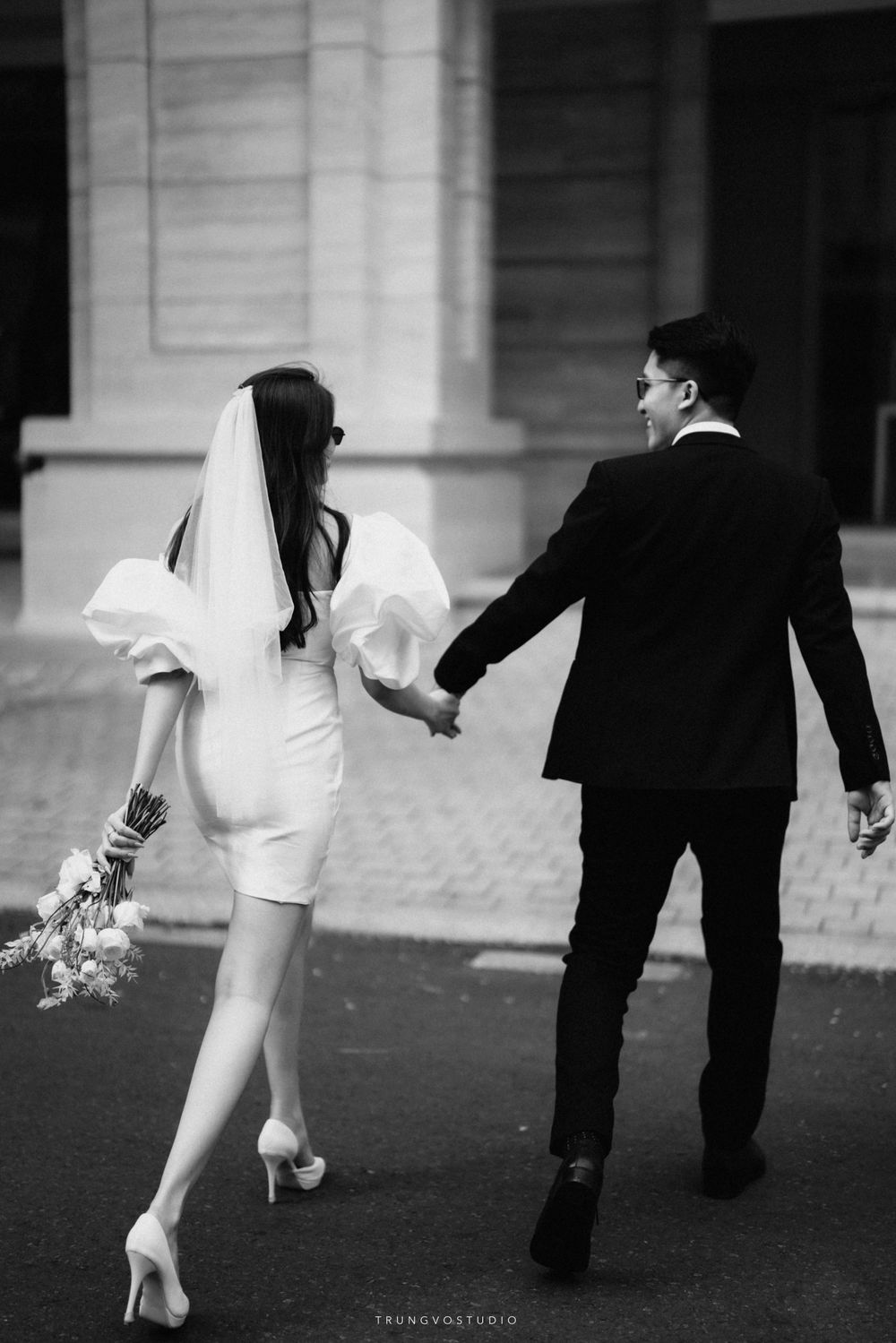 Bride in short white dress and groom in black suit walk hand in hand in a black and white wedding photo.
