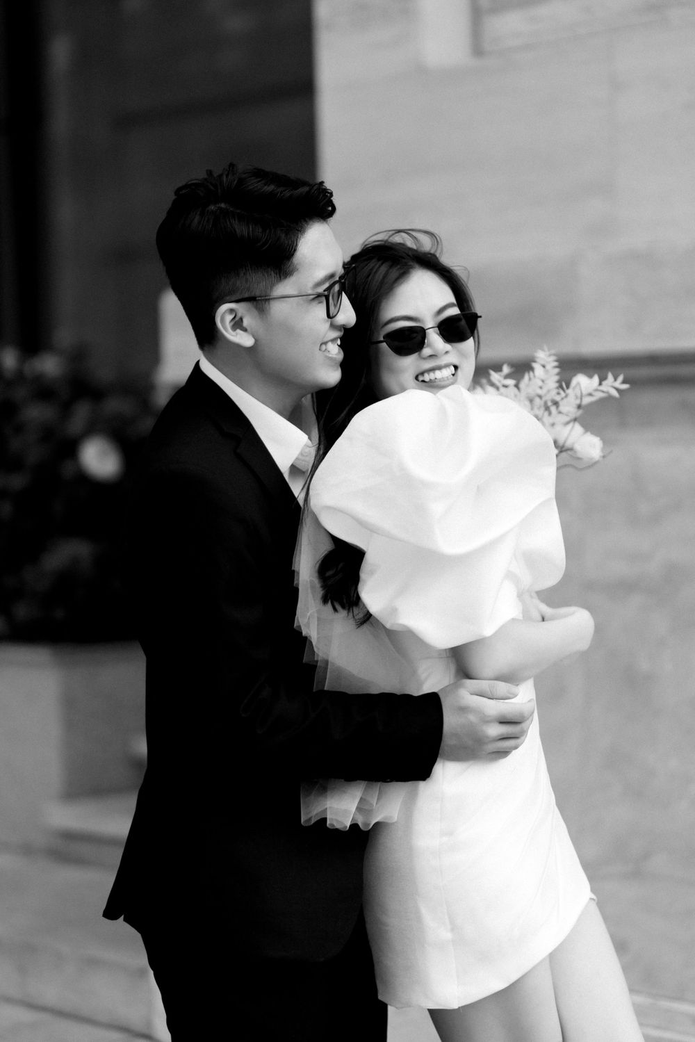Groom in black suit embraces smiling bride in white ruffled mini dress and sunglasses outdoors.