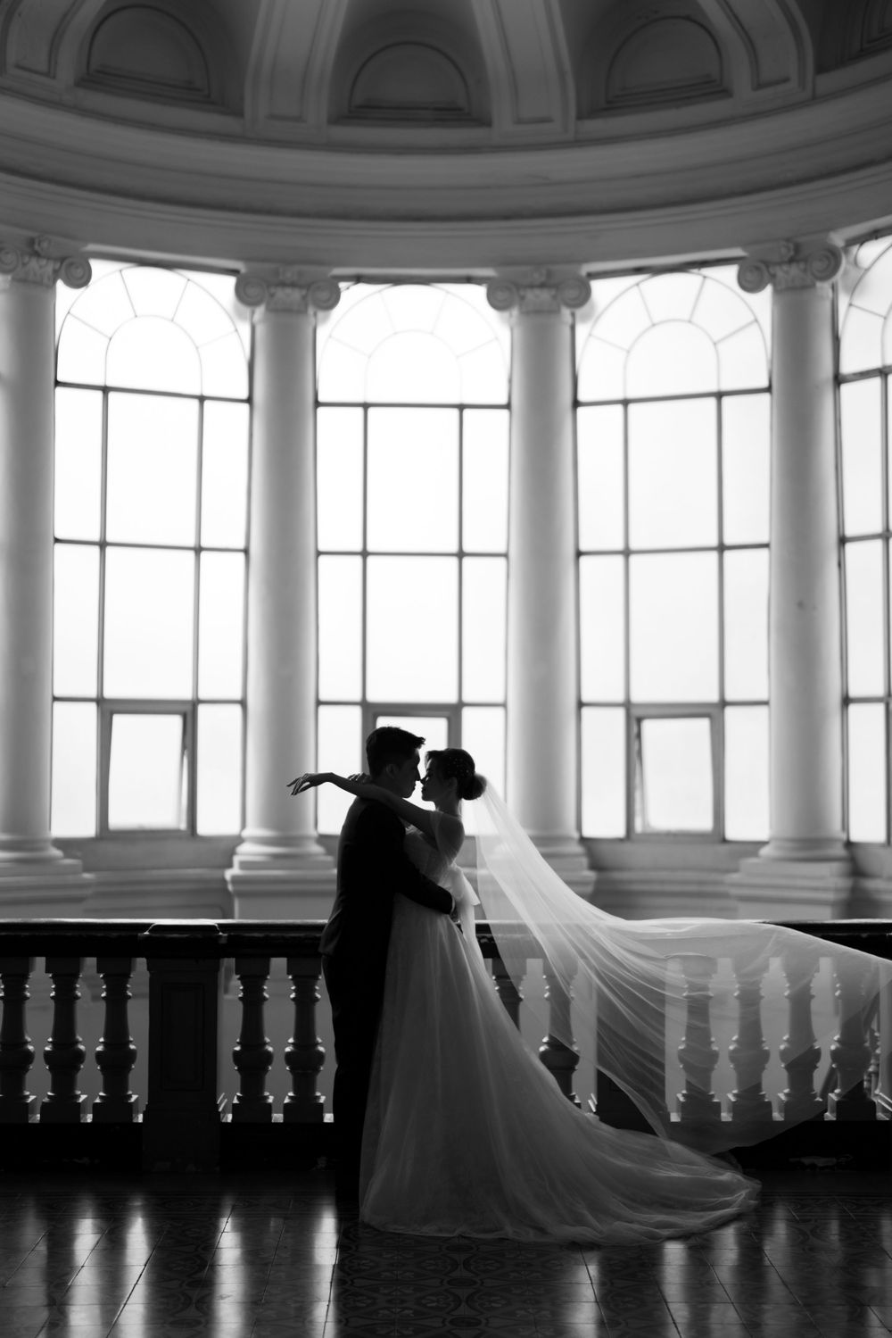 Bride and groom silhouetted in romantic embrace before arched windows with flowing veil in grand hall.