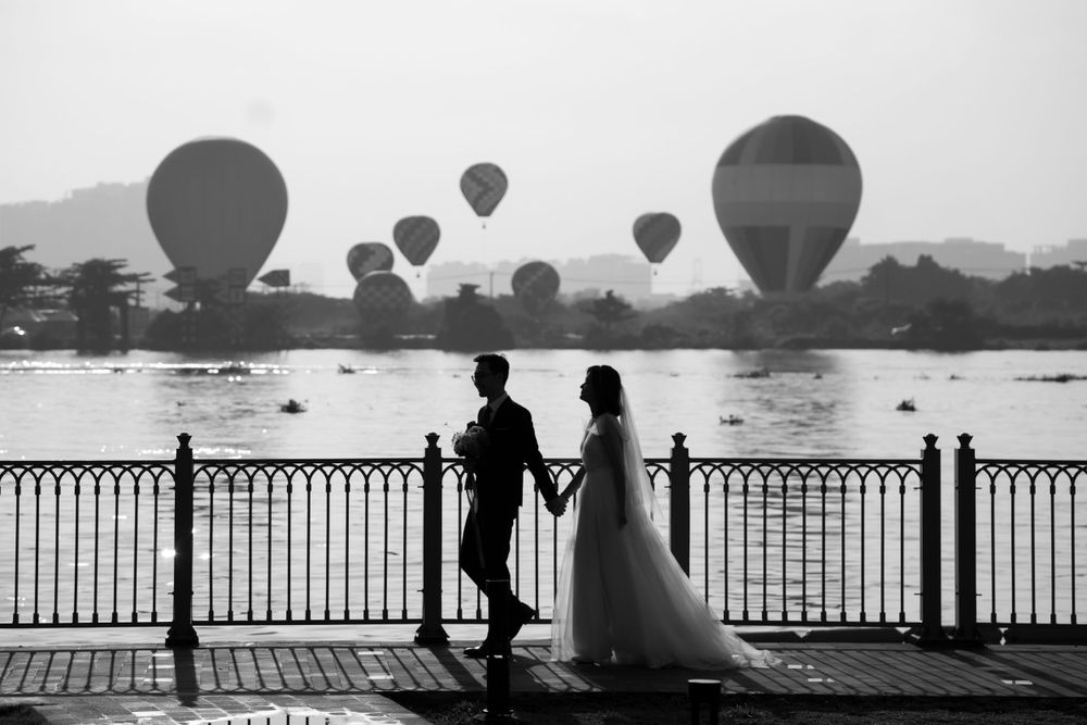 Bride and groom silhouetted on waterfront bridge with hot air balloons floating in misty sky behind them.