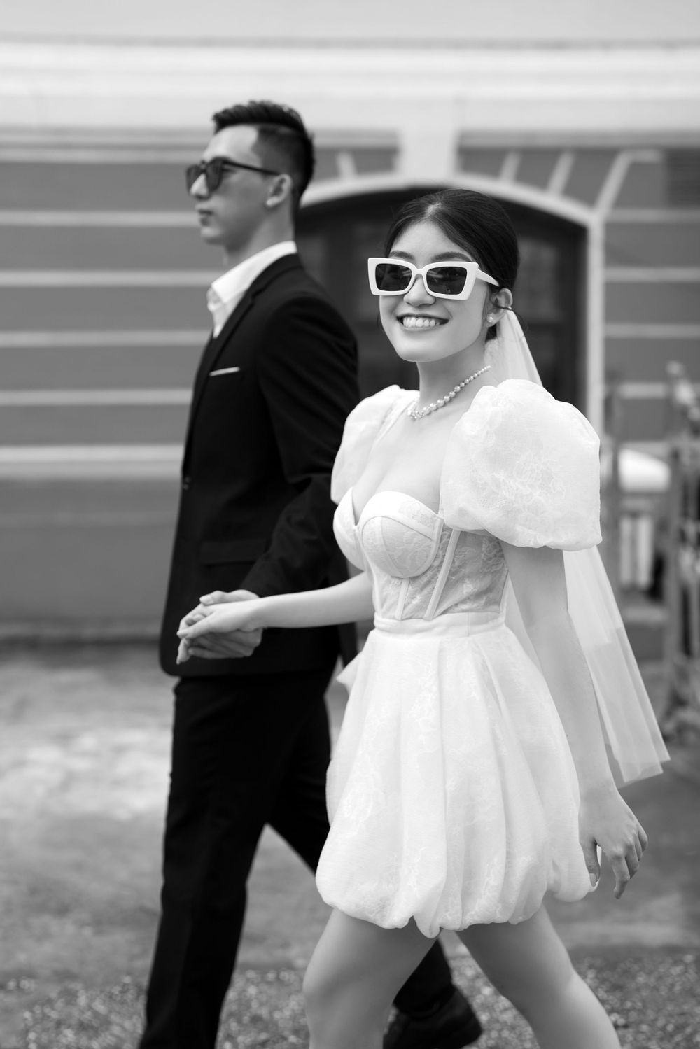 Smiling bride in white mini dress and sunglasses leads groom in black suit by the hand outdoors.