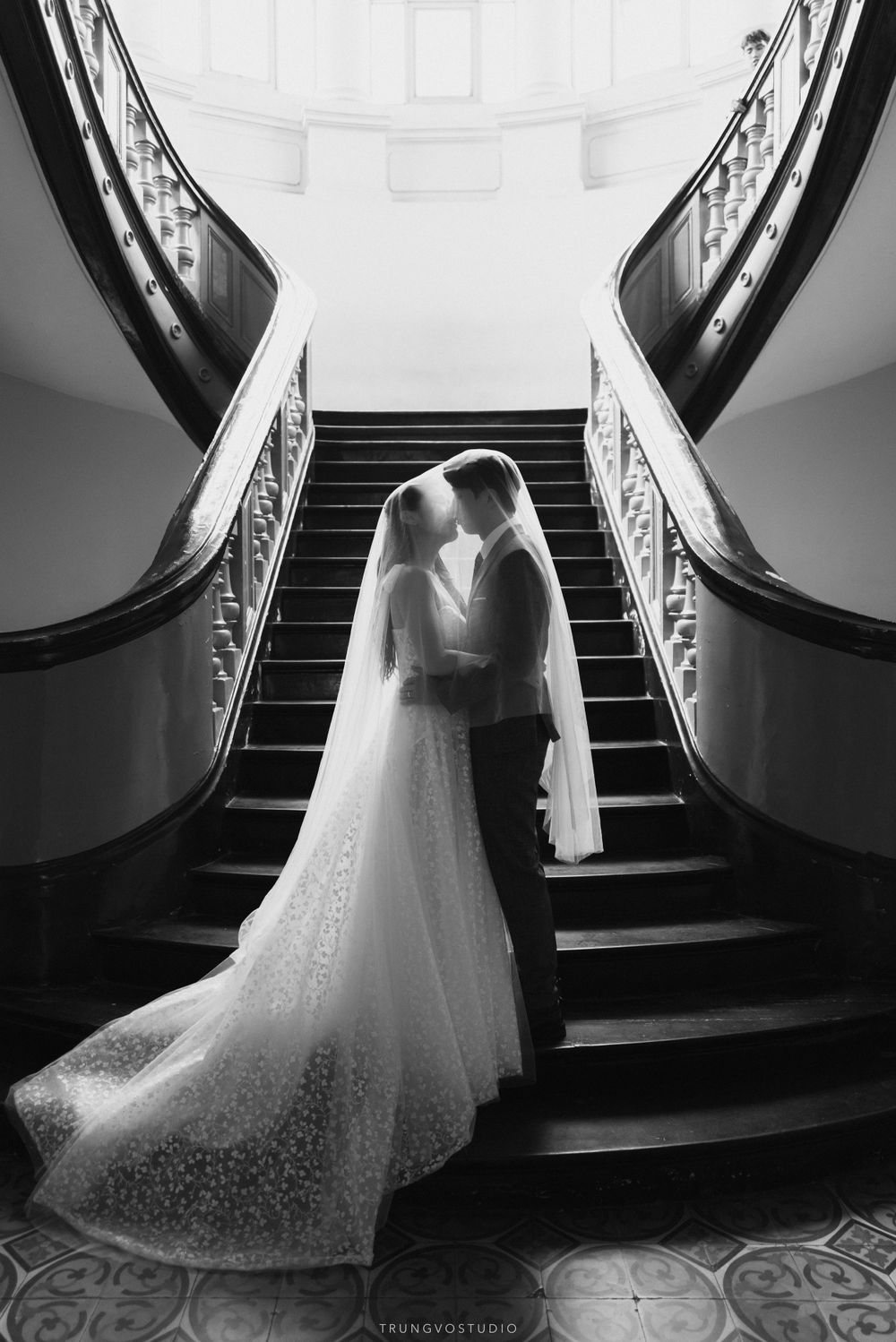 Bride and groom share intimate moment under veil on grand staircase in stunning black and white photo.