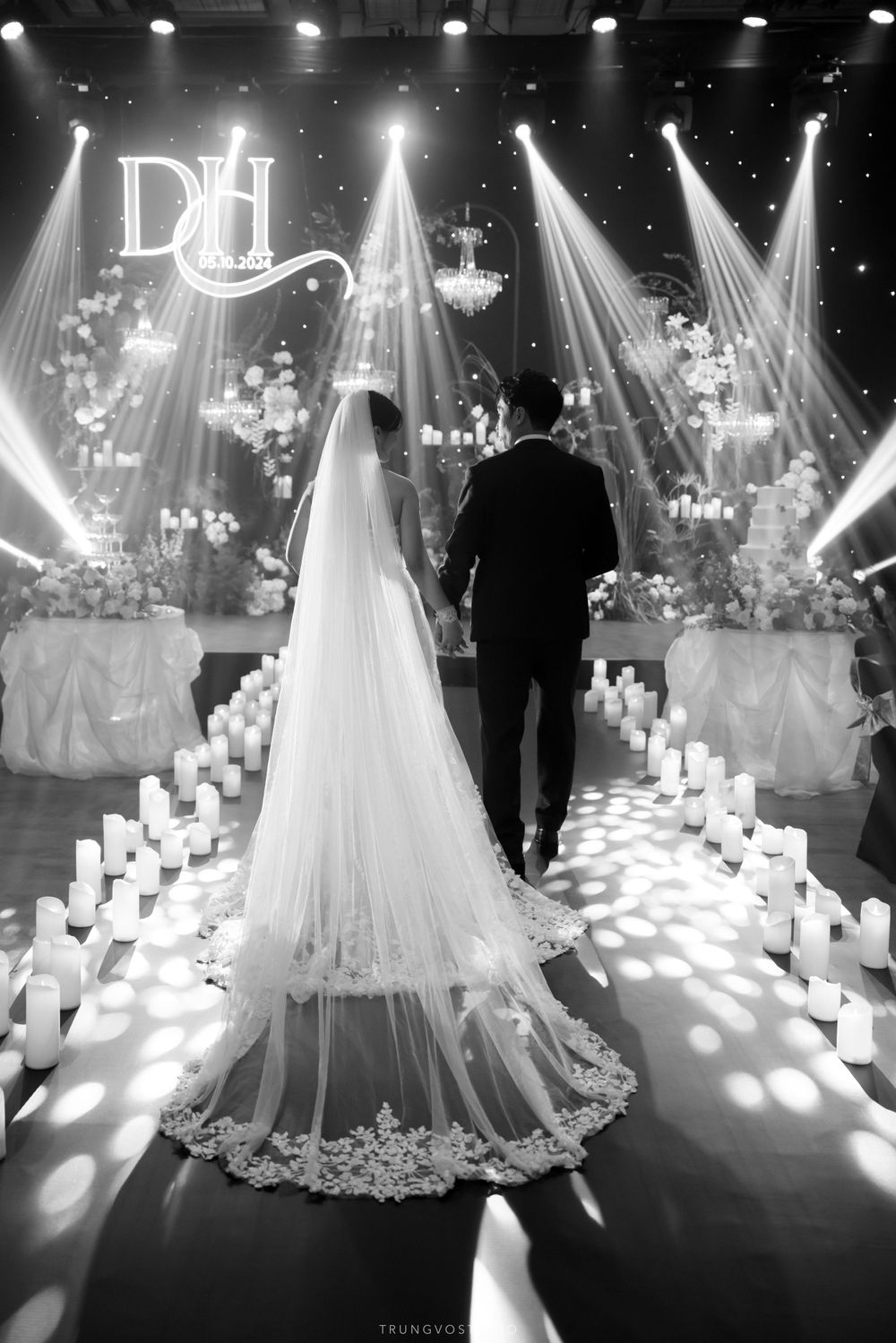Bride and groom walk down candlelit aisle toward illuminated floral wedding stage in black and white.