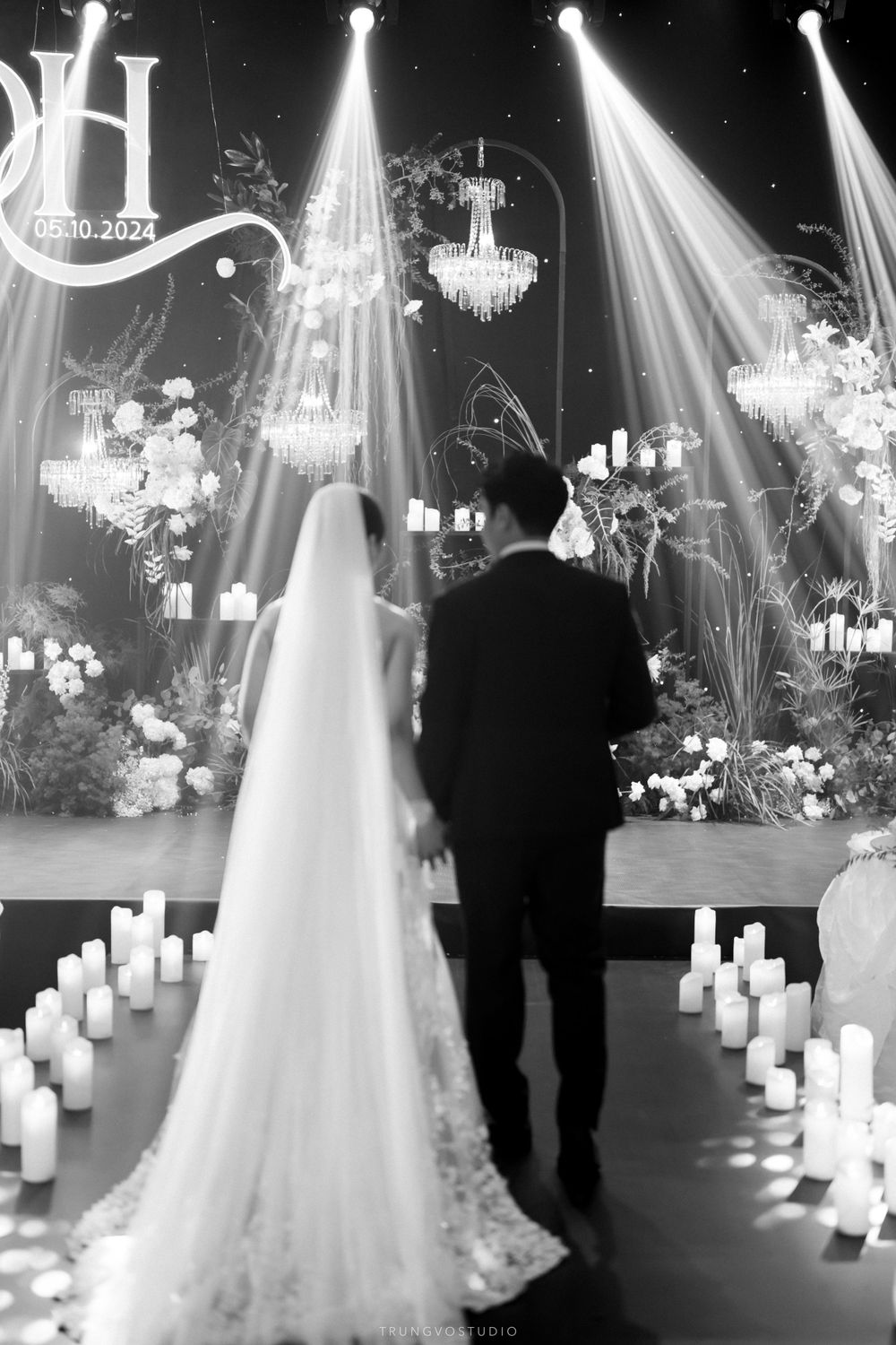Black and white wedding photo of bride and groom facing floral altar with candles and chandeliers.