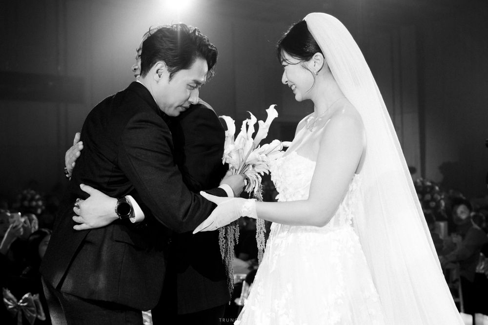 Black and white photo of groom embracing emotional bride holding white flowers at wedding ceremony.
