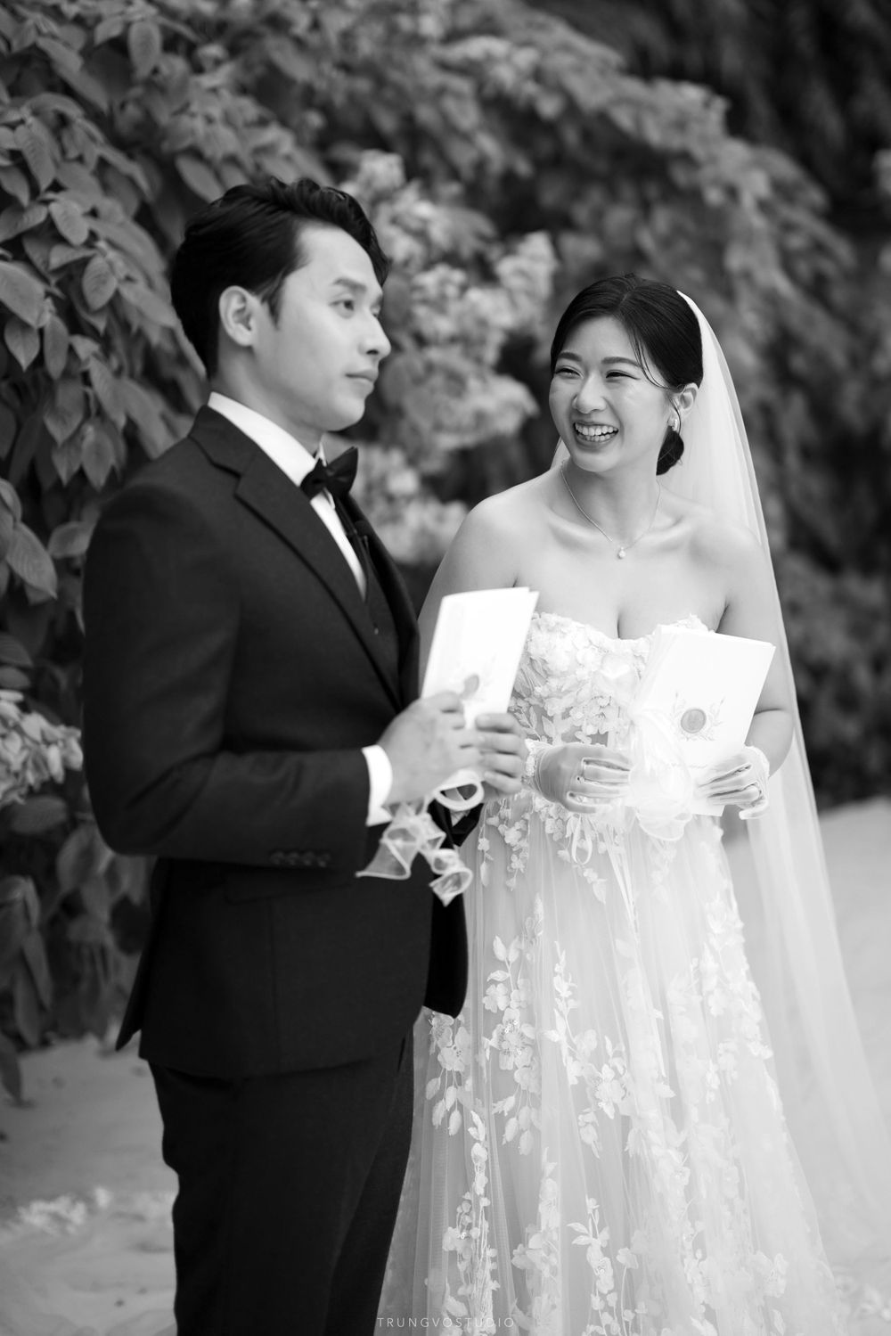 Bride in lace gown and groom in tuxedo laugh together while reading wedding vows outdoors.