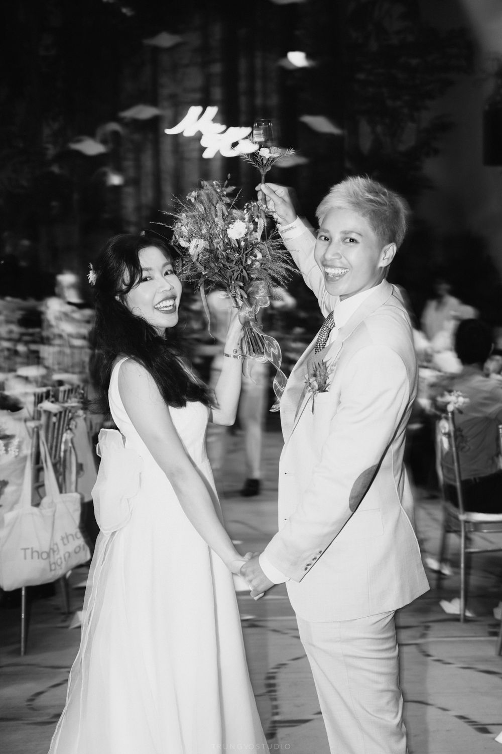 Two brides hold hands and smile, one in a white dress holding a bouquet aloft at their wedding reception.