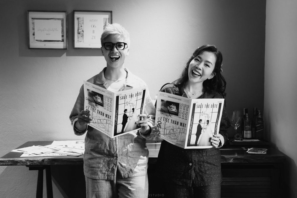 Couple smiling and holding wedding newspapers in a black and white indoor photo.