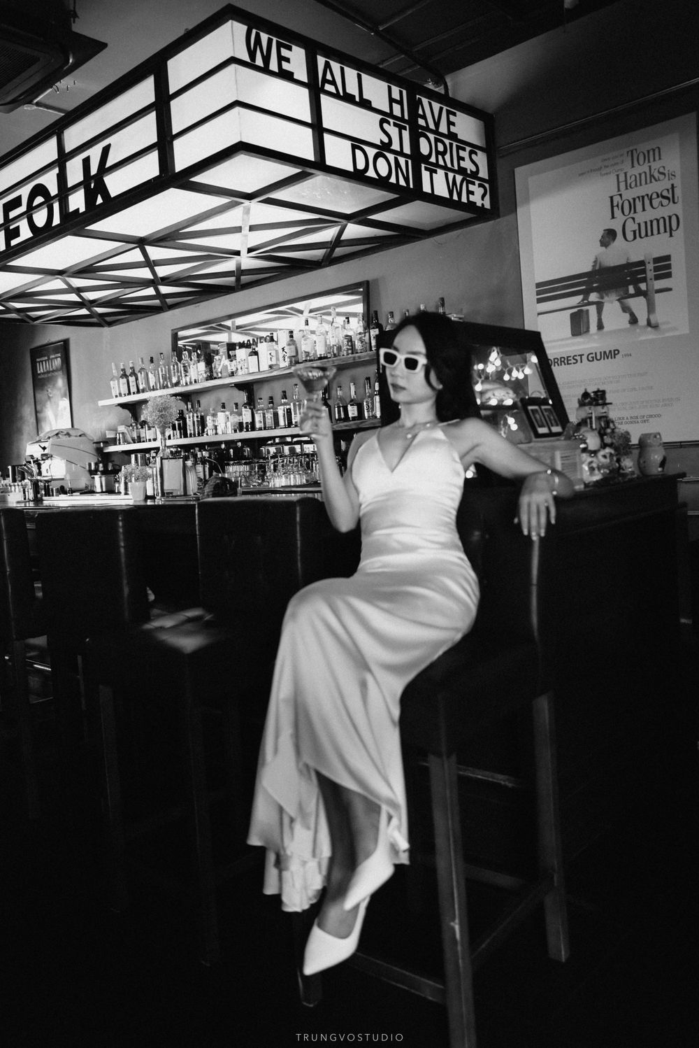Black and white photo of bride in white dress posing at bar with sunglasses.