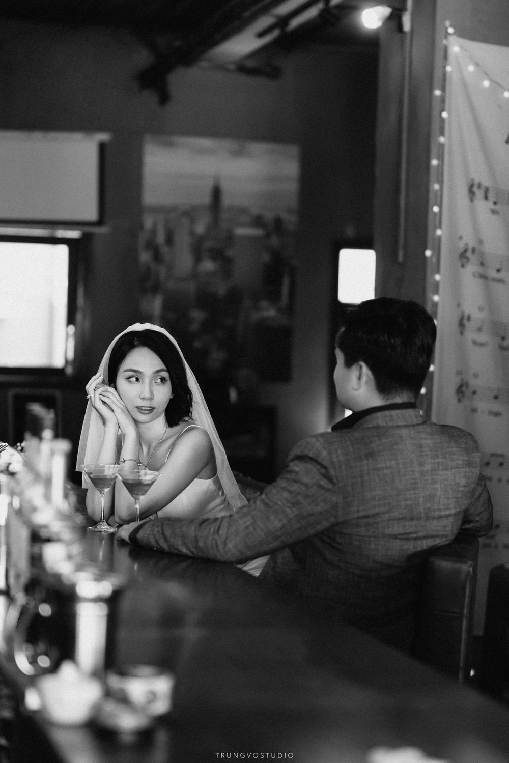 Black and white photo of bride in veil gazing at camera while seated at bar with groom.