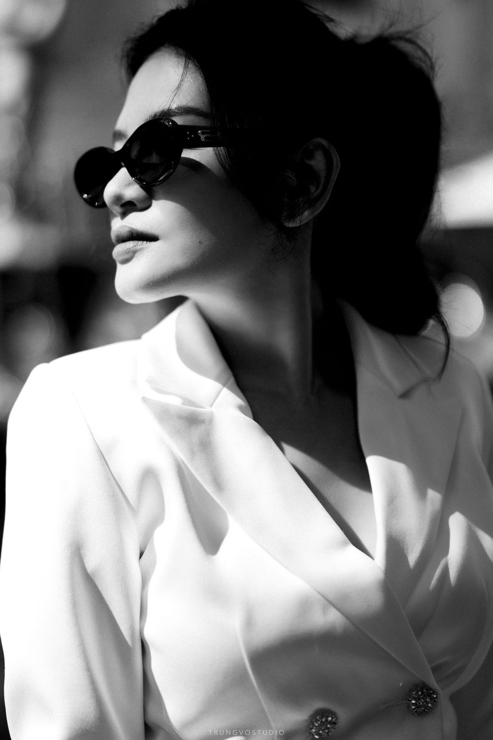 Elegant black and white portrait of woman in white blazer wearing sunglasses in sunlight.