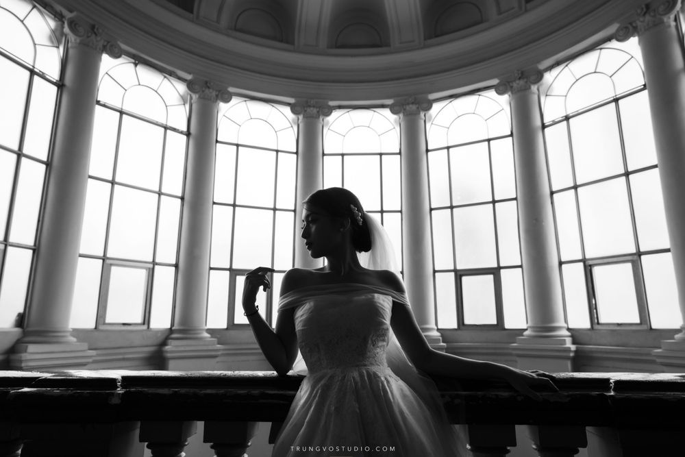 Bride in off-shoulder gown stands before arched windows in black and white silhouette portrait.