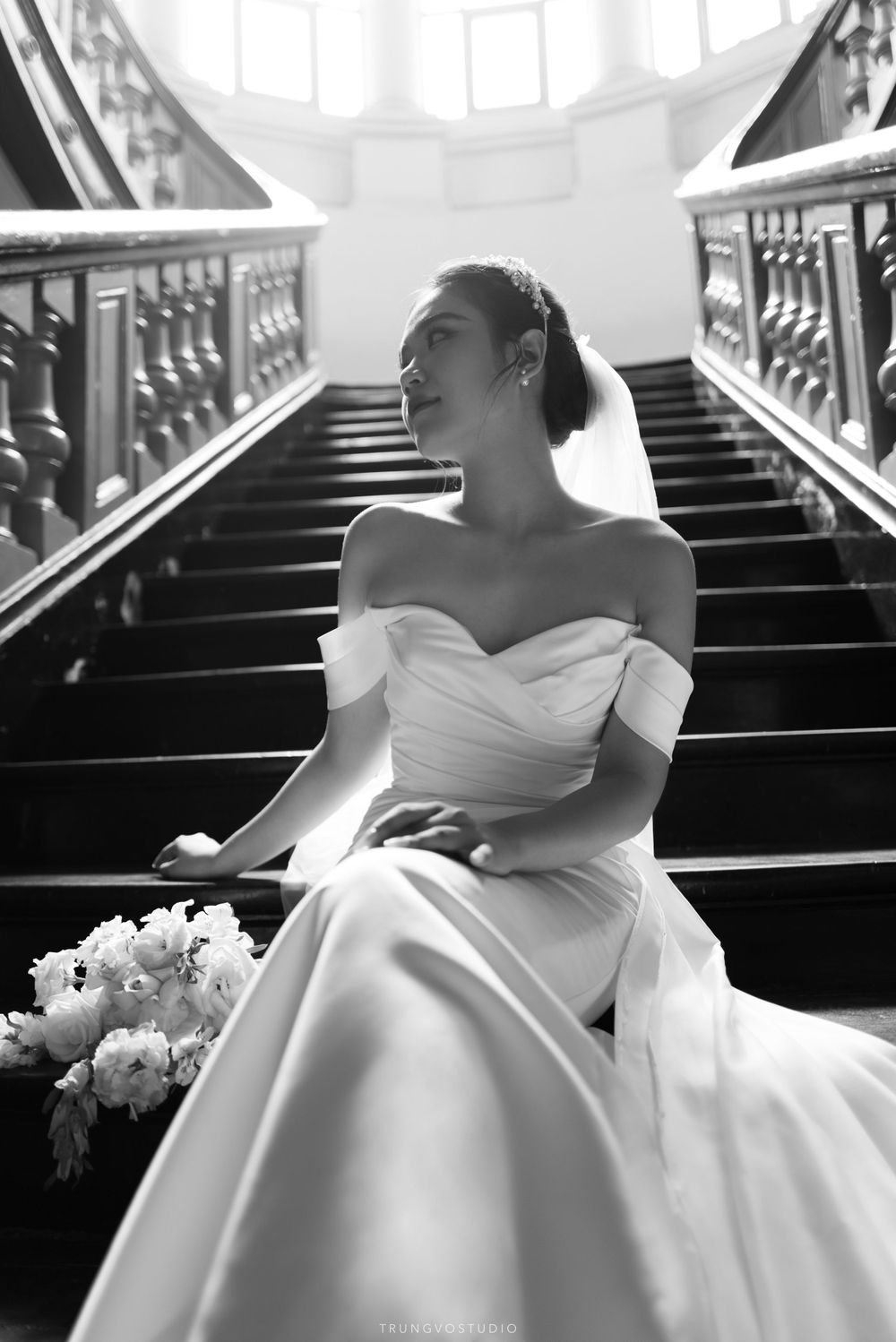 Bride in off-shoulder gown sits on grand staircase holding bouquet, black and white wedding portrait.