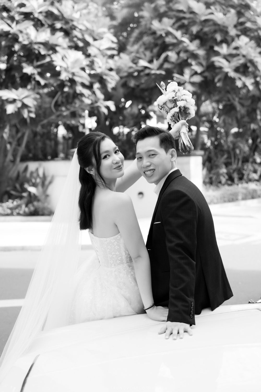 Bride and groom pose together beside vintage white car, black and white wedding photo outdoors.