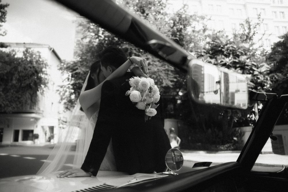 Bride and groom reflected in vintage car window holding bouquet, artistic black and white photo.