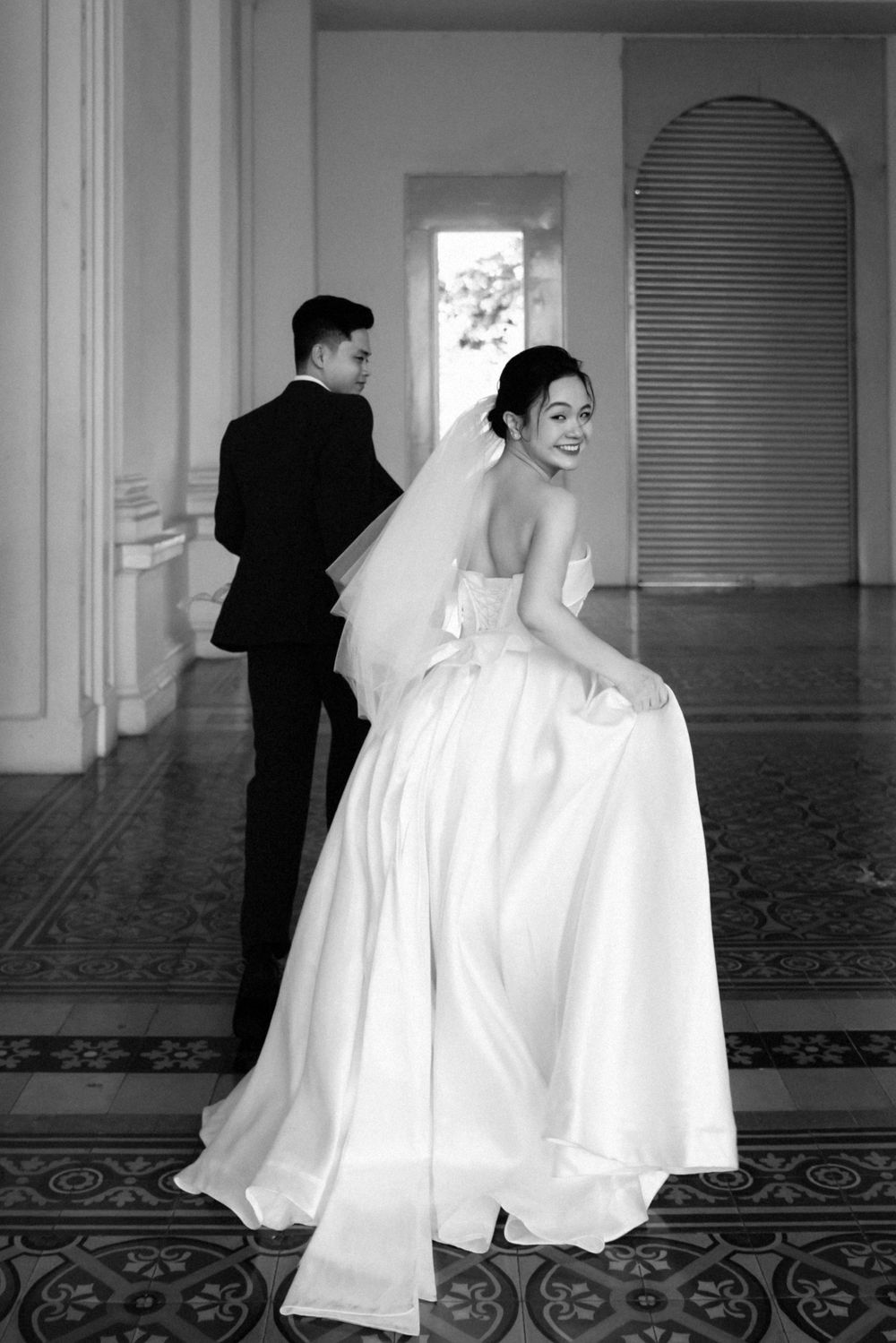 Bride in strapless gown glances back smiling while groom walks ahead in elegant hallway portrait.