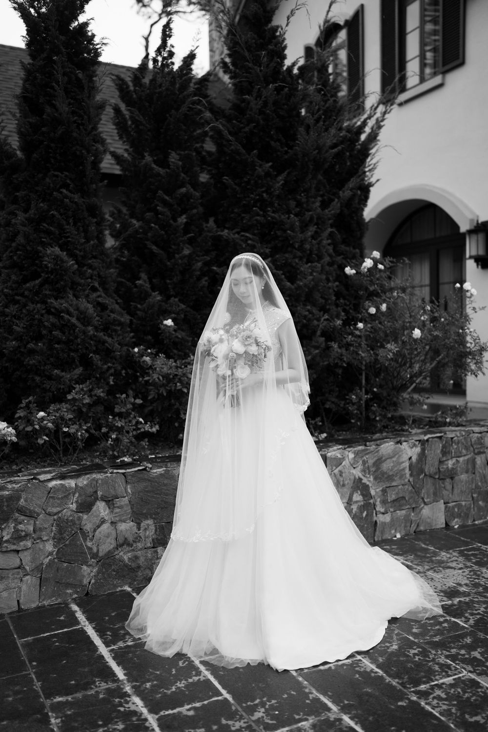 Bride in full veil and flowing gown holds bouquet before trees and stone building, black and white.