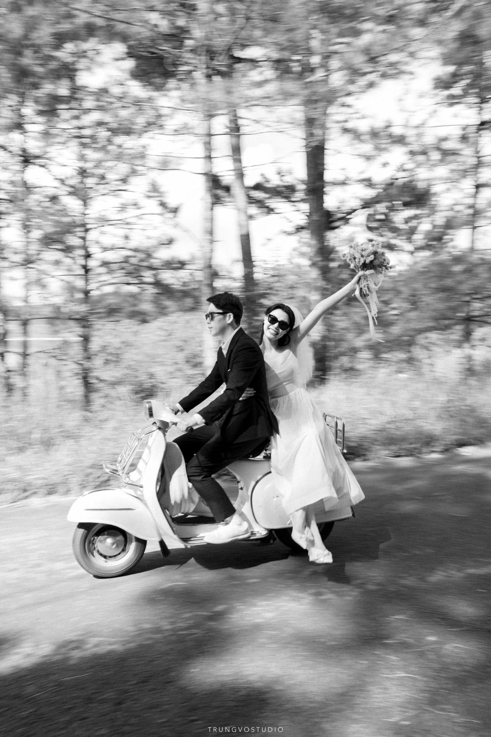 Bride and groom ride Vespa scooter through forest road, bride raising bouquet, black and white.