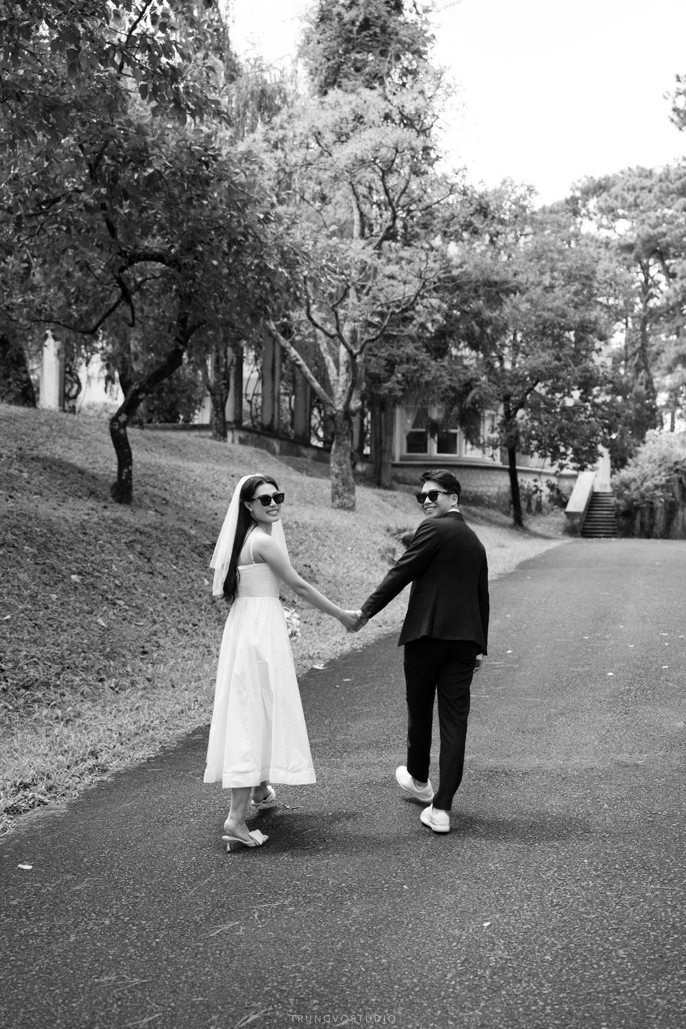 Bride and groom hold hands walking down tree-lined path wearing sunglasses, black and white photo.