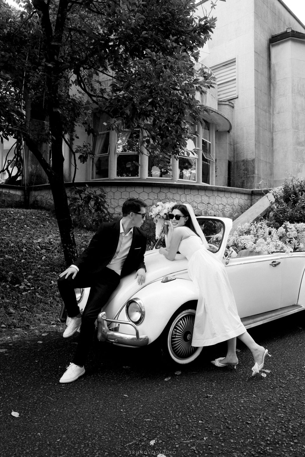 Bride and groom leaning against a vintage white Volkswagen Beetle in a romantic black and white wedding photo.