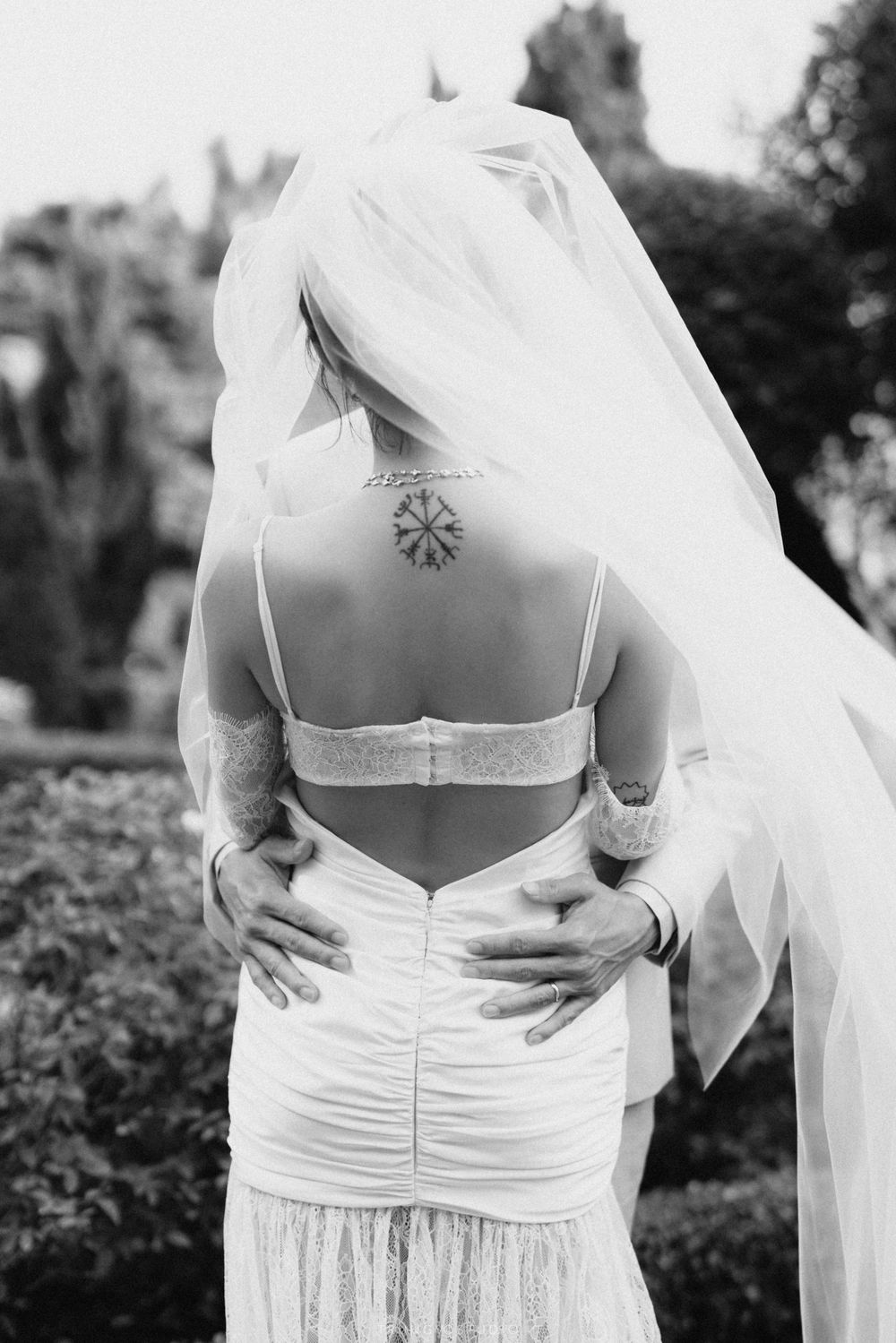 Black and white photo of a bride showing her tattooed back in a backless wedding gown with a flowing veil.