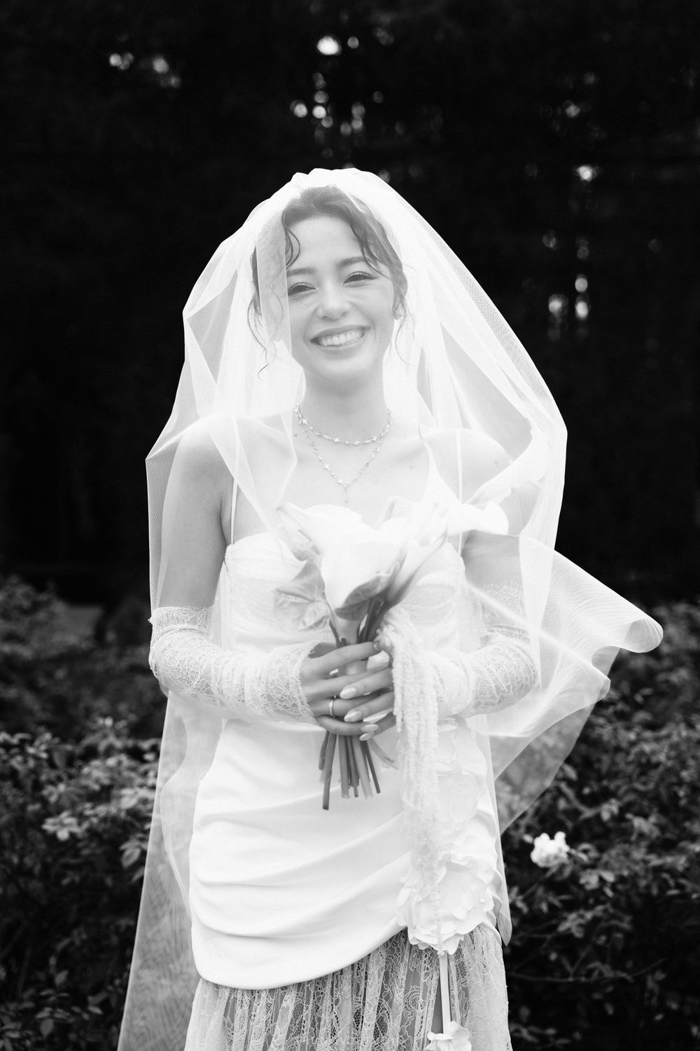 Smiling bride in a white wedding dress and veil holding a bouquet in a black and white outdoor portrait.