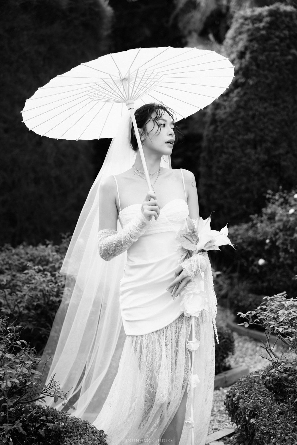 Elegant bride in a white wedding gown holding a parasol and floral bouquet in a lush garden setting.