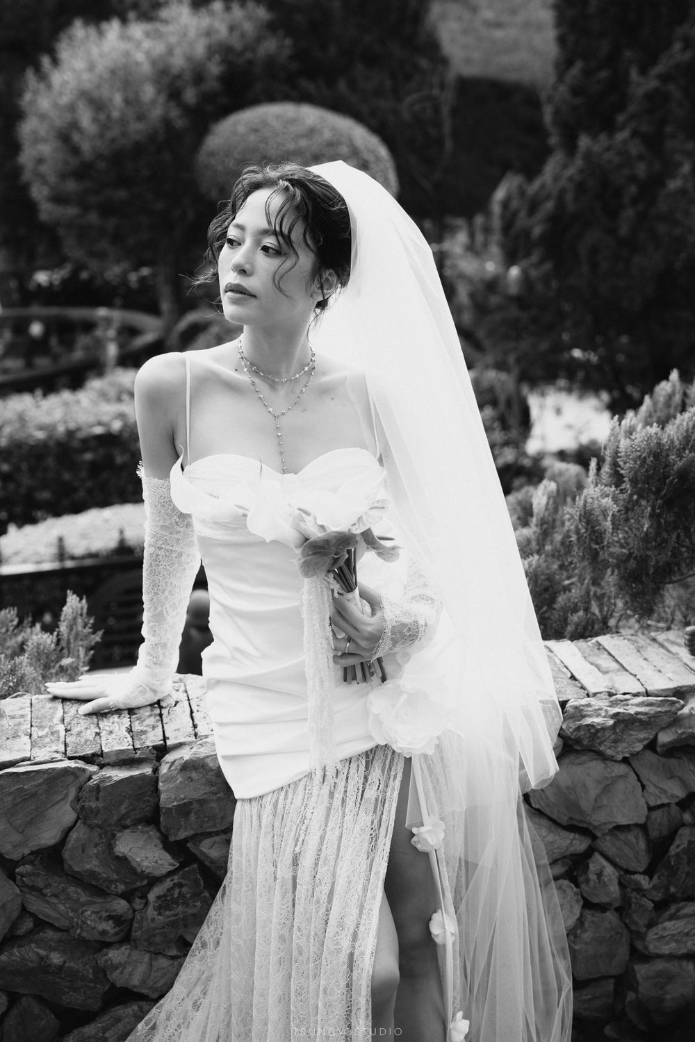 Bride in a strapless white wedding dress with veil and floral bouquet posing by a stone wall in a garden.