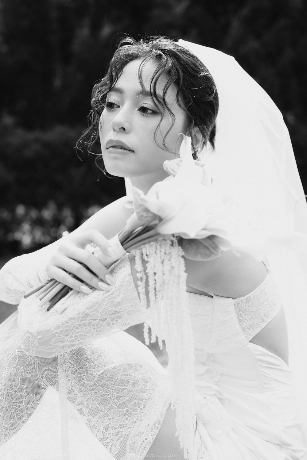 Elegant bride in a lace wedding dress and veil holding a bouquet in a contemplative black and white portrait.