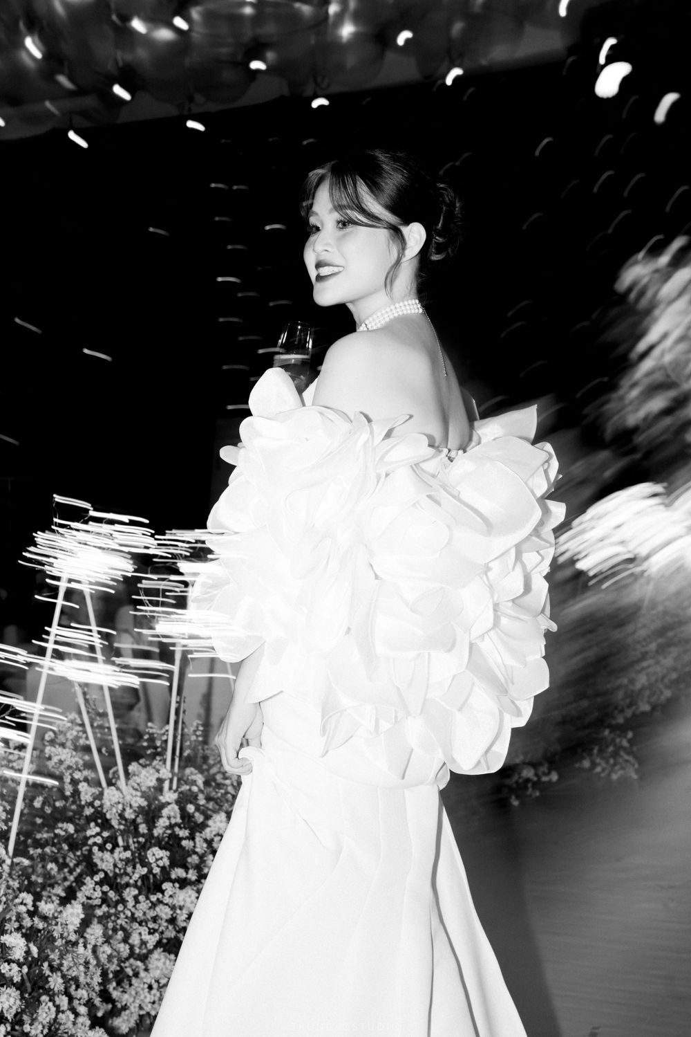 Bride in a stunning off-shoulder ruffled white gown posing at a floral-decorated wedding reception venue.
