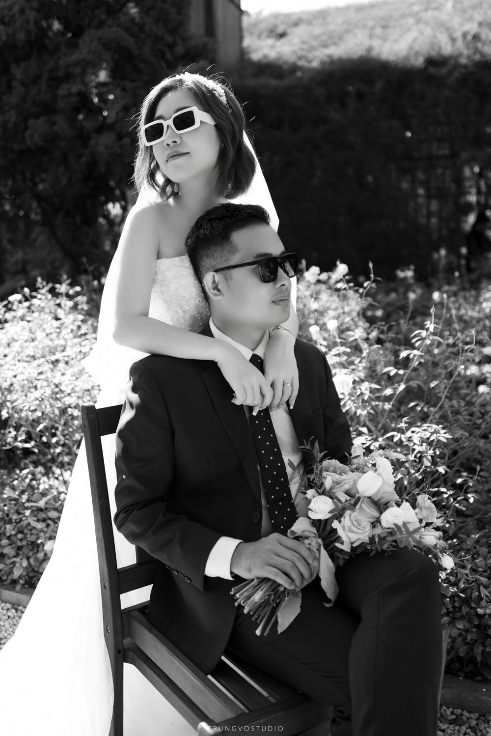 Black and white photo of a stylish bride and groom wearing sunglasses in a garden setting.