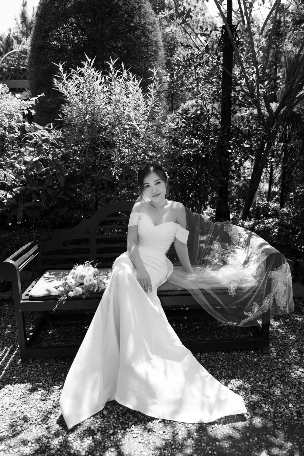 Black and white photo of a bride in an off-shoulder gown sitting on a bench in a garden.