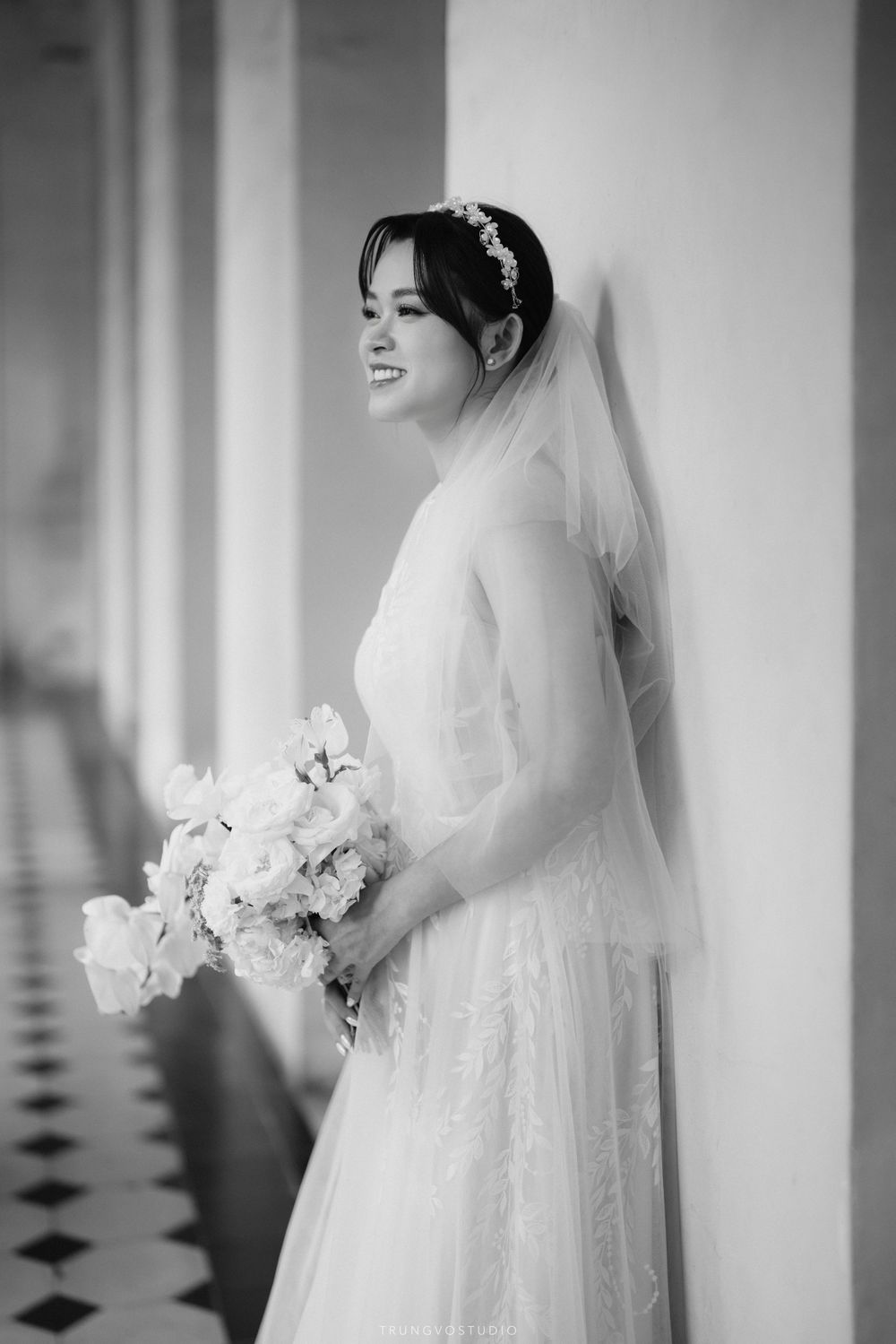 Black and white photo of a smiling bride holding a bouquet while leaning against a wall.