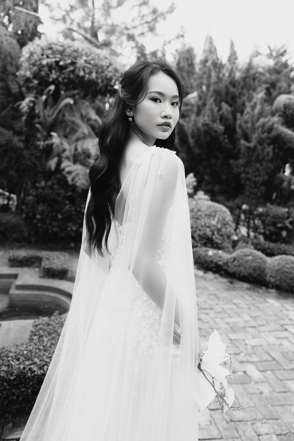 Black and white photo of Asian bride in white gown with flowing cape in garden landscape.