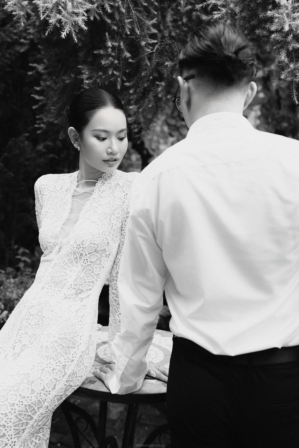Bride in white lace dress looking down at groom in casual white shirt, intimate black and white portrait.