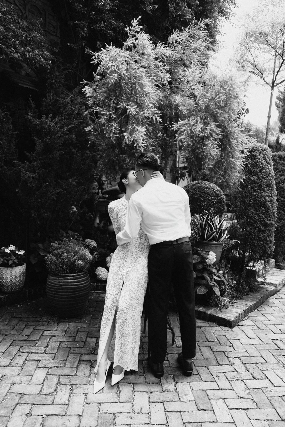 Black and white photo of bride and groom embracing in a lush garden courtyard on their wedding day.