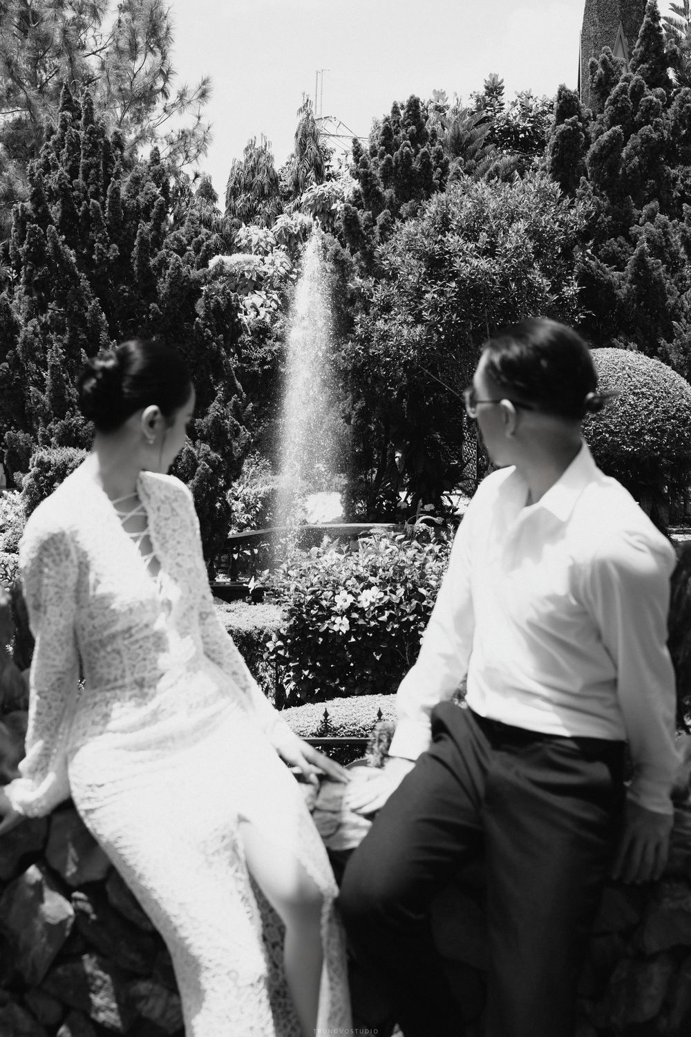 Black and white wedding photo of bride and groom sitting by a garden fountain sharing a quiet moment.