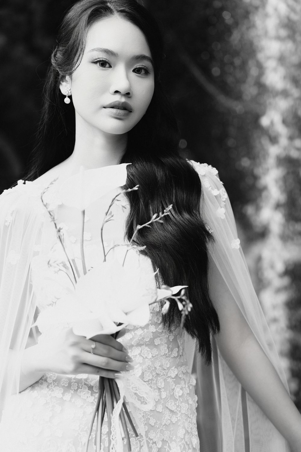 Black and white portrait of Asian bride holding white flowers wearing lace gown with sheer cape.